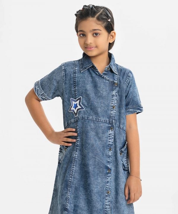 Kids girl A-line retro wrap style tunic in denim fabric. Classic collar, Short sleeved. Button fastening at the front. inseam pockets and attached patch at the front.